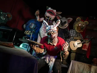 Le personnage de Traumatica « Serveur zombie » et cinq monstres en peluche Personnage de Traumatica : serveur zombie avec une coiffe en forme de tête de lièvre et un plateau en main. En arrière-plan, un gigantesque monstre en peluche avec de longues dents