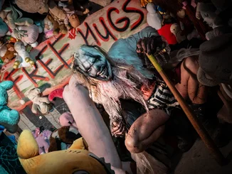 Clown de Traumatica « Câlins Gratuits » Personnage de Traumatica avec tête de clown affreuse qui se tient devnat un mur plein de poupées et d’animaux en peluche. Au milieu un panneau avec l’inscription « Free Hugs » (câlins gratuits).
