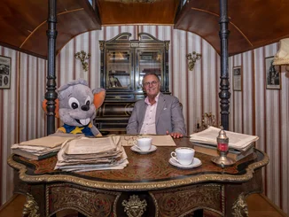 Roland Mack sits at a desk in a historic circus wagon with a large Ed soft toy.