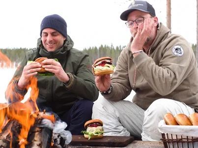 Thomas Mack and Brian Bojsen are sitting together in the snow by a fire, eating burgers.
