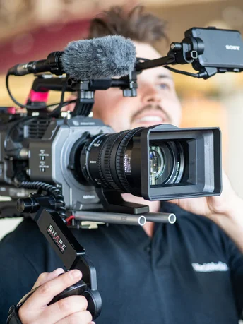 Ein Videograf von MACK Media mit einer Kamera auf der Schulter schaut lächelnd durch den Viewfinder der Kamera.