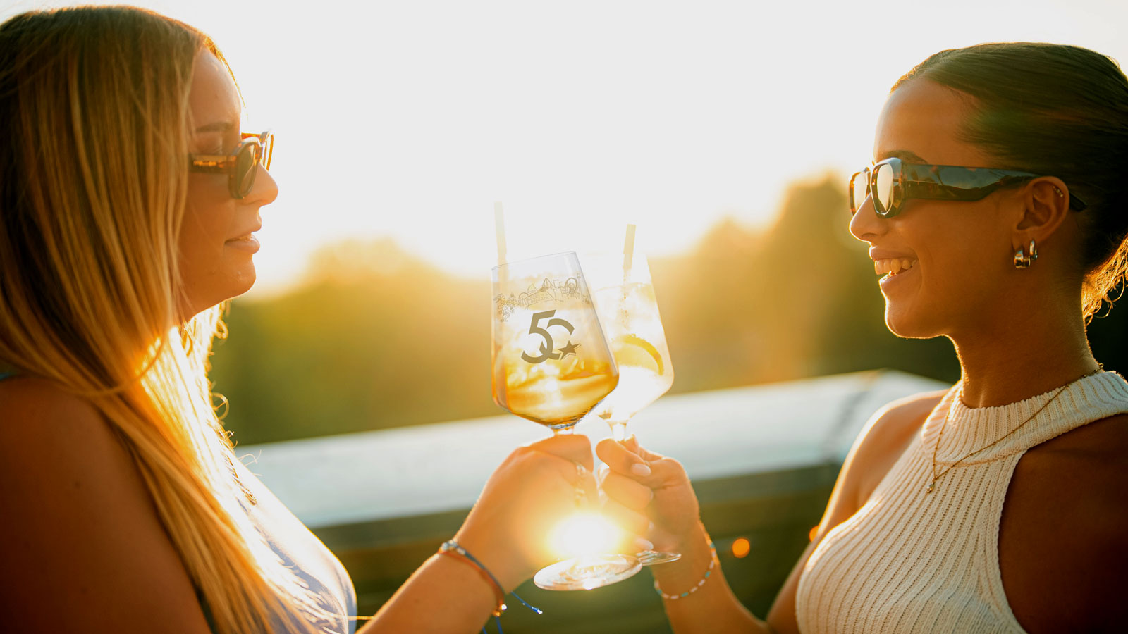 Zwei junge Damen trinken im Sonnenuntergang aus den Jubil&auml;ums-Weingl&auml;sern. 