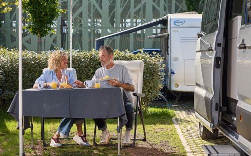 A couple are having breakfast at a camping table next to their caravan on a caravan pitch at the Europa-Park Resort.