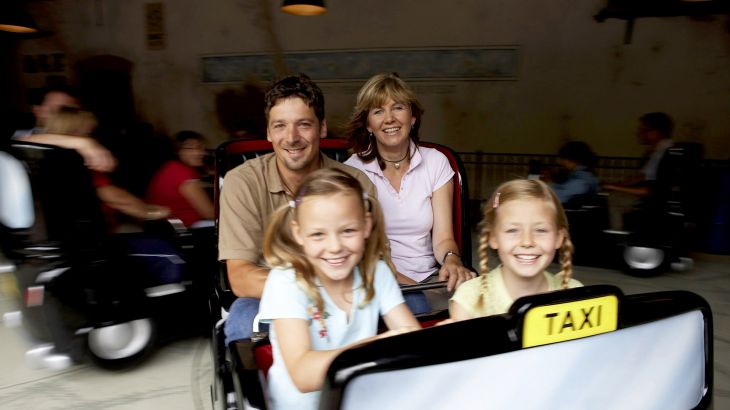 Ein Paar und zwei Kinder sitzen lachend im Crazy Taxi im Englischen Themenbereich des Europa-Park. Im Hintergrund sieht man weitere Fahrgäste.