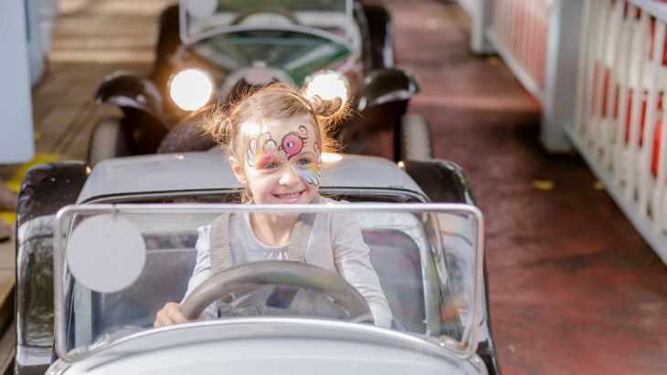 A girl with face paint is sitting in a vintage car at the Lada Autodrom in the Russian themed area of Europa-Park, smiling.