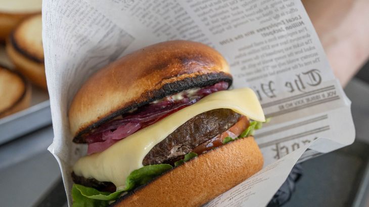 A close-up of a juicy cheeseburger at the Gin Tonic & Burger event at the ‘Bell Rock’ hotel. The burger is wrapped in a paper bag with a newspaper print.