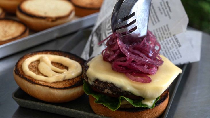 A close-up of a cheeseburger being topped with red onions. In the background are a paper bag with a newspaper print and some more burger patties.