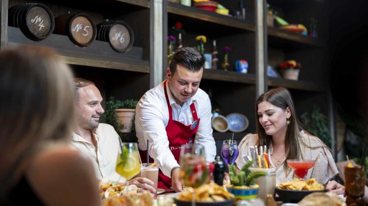 At the Taco Fiesta, a waiter wearing a red apron serves Mexican specialities such as tacos, wraps and burgers, and colourful cocktails are also on the table. Guests laugh and enjoy the food together.