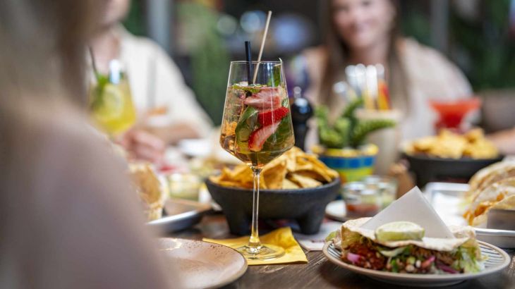 A Mexican cocktail with fresh strawberries is surrounded by typical Mexican dishes such as tacos, nachos and wraps on a decorated table.