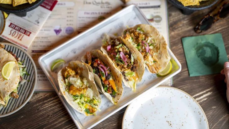 Four freshly prepared Mexican-style tacos with fresh ingredients, served on a table with crispy nachos.