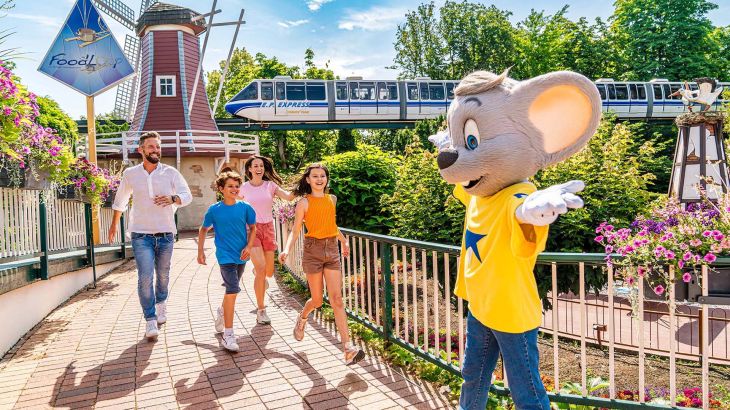 Une famille en habits estivaux marche tout sourire en direction d’Ed Euromaus sur le pont devant le restaurant FoodLoop à Europa-Park. L’EP-Express circule en arrière-plan.