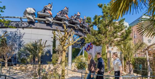 Drei Europa-Park Besucher laufen durch den Wartebereich der Silver Star im Themenbereich Monaco. Dort stehen viele Bäume. Im Hintergrund sieht man mehrere Wagen der Silver Star mit Fahrgästen.